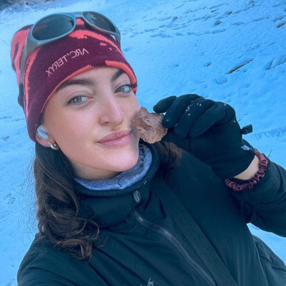 Person in winter clothing eating a piece of Campers Pantry freeze dried chocolate ice cream perfect for hiking and adventures against a snowy background