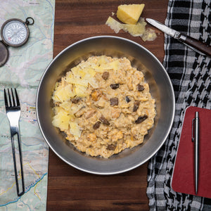 A large silver bowl filled with a Campers Pantry creamy chicken and mushroom pasta hiking food meal, surrounded by map-related items and a piece of cheese on a napkin.