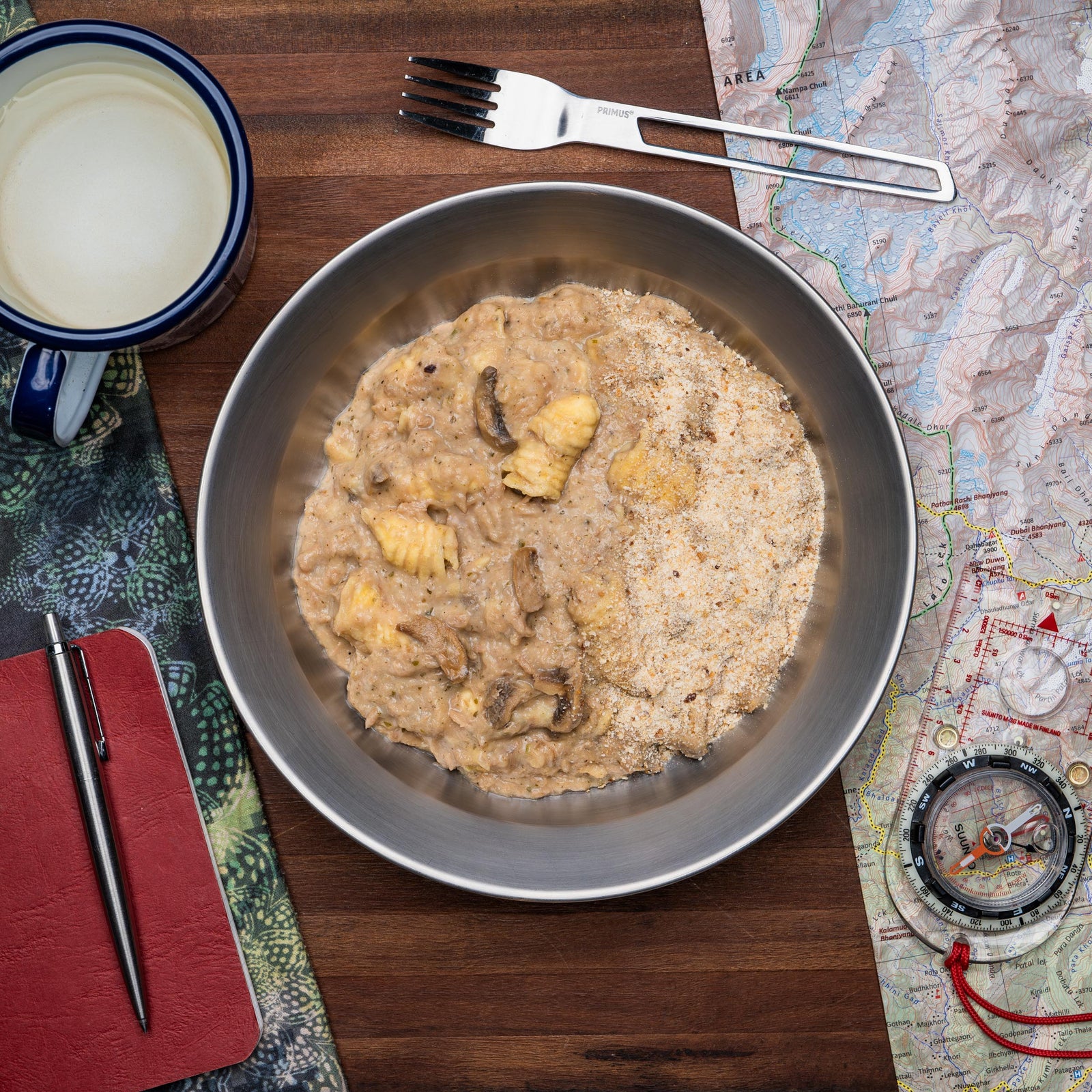 A Campers Pantry tuna pasta bake hiking food meal in a metal bowl, with a fork and knife on top of the bowl, surrounded by a map, a compass, and a notebook. Using real ingredients, these meals are fully cooked before drying and packing.