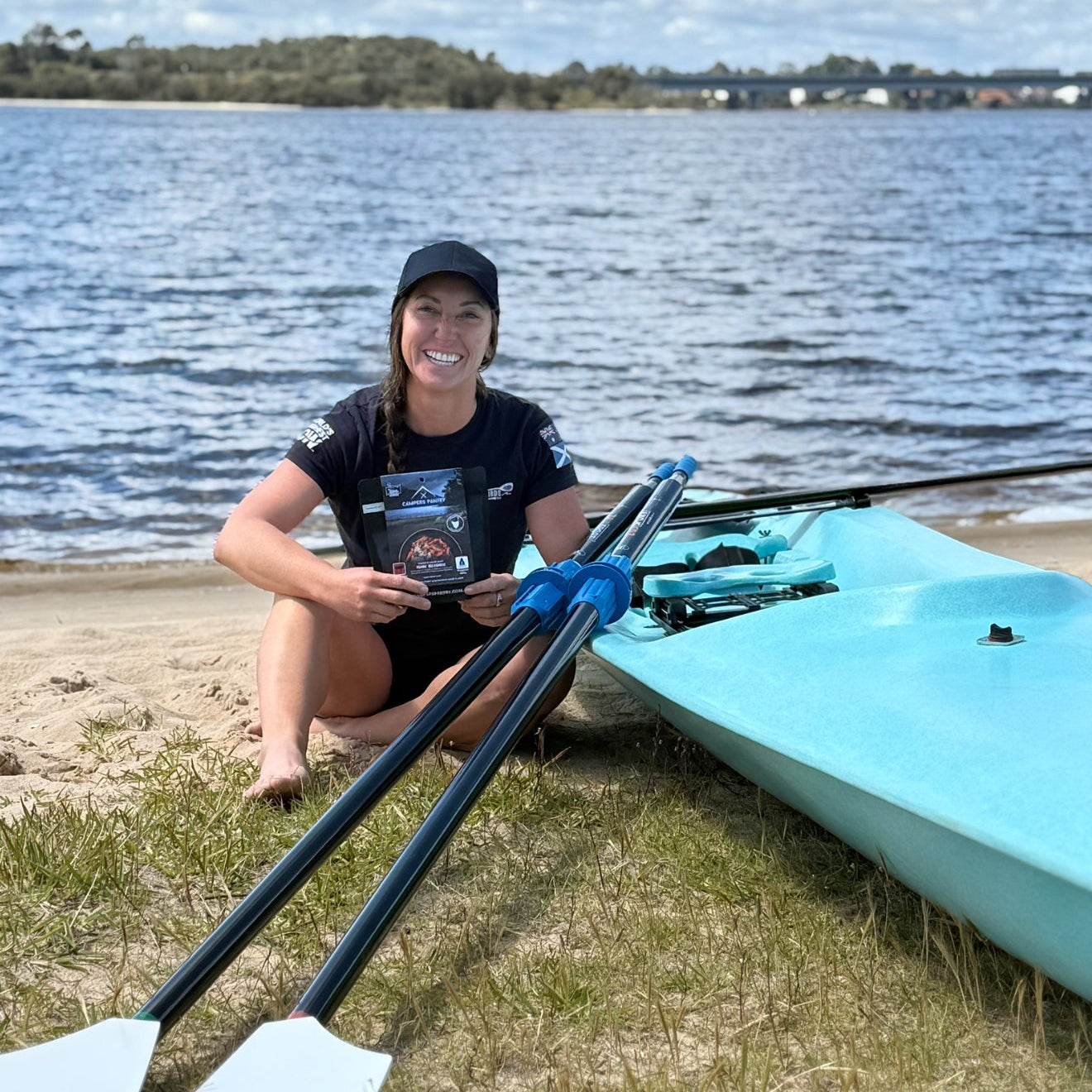 Karla Pound - Team Atlantic Warterbirds, sitting on a beach with a rowing boat and oars, smiling at the camera holding a packet of Campers Pantry freeze dried hiking food.