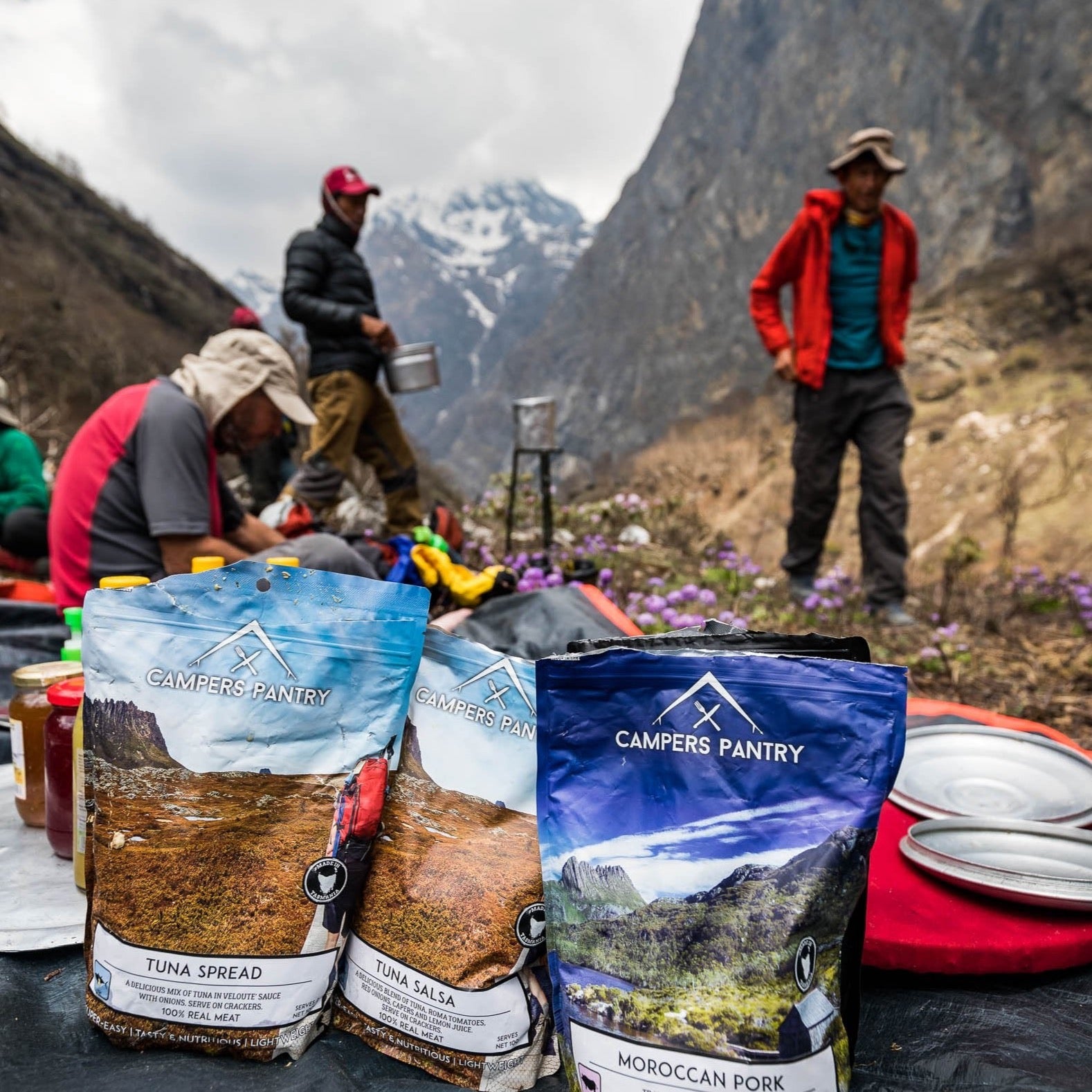 Packaged camping food from Campers Pantry displayed in a outdoor setting with individuals in the background engaging in camping activities. Hiking food which is lighter then dehydrated meals.
